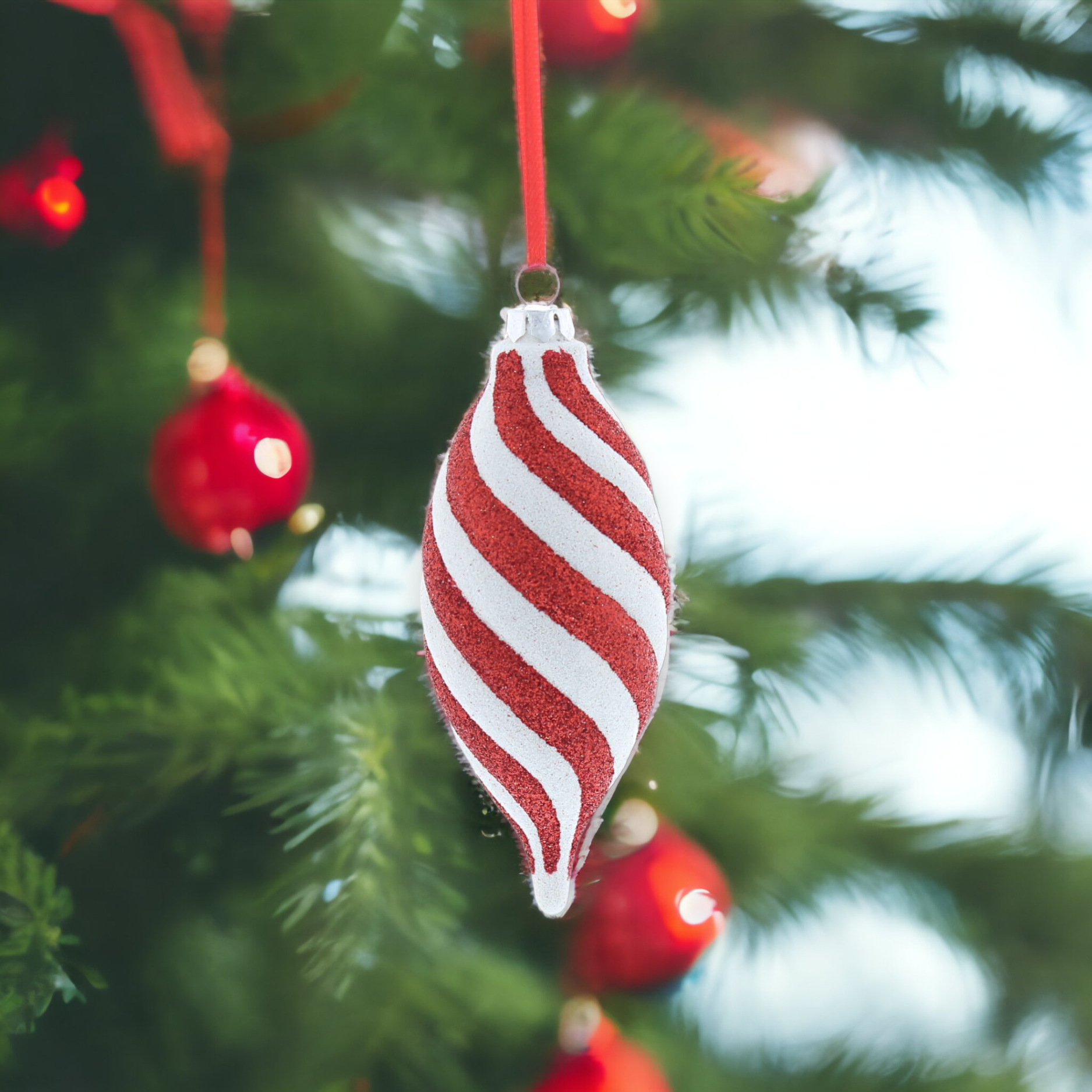 A red and white striped glitter hanging decoration resembling a candy, placed on a Christmas tree with red ornaments and green leaves.