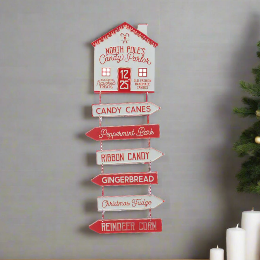A tall, vertical, red and white sign with directional letters and candy names, placed against a backdrop of a real Christmas tree.