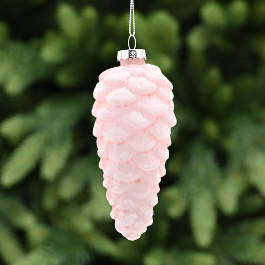 A light pink frosted glass pinecone Christmas tree decoration with white glitter, hanging from a silver wire, displayed against a blurred green pine background.