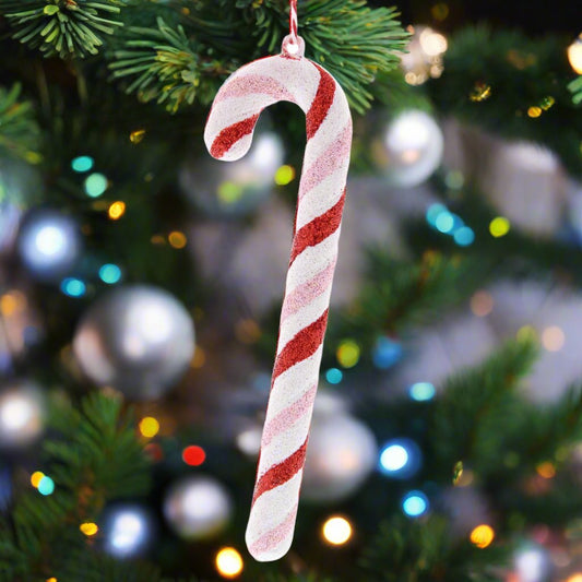 A hanging decoration featuring a candy cane with pink and red stripes, with a blurred Christmas tree and lights in the background.