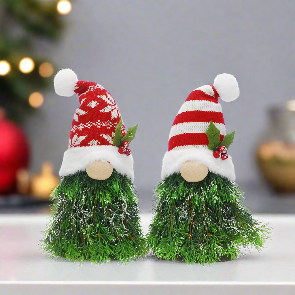 Two decorative Christmas tree gonks with snowflake and striped hats, made of fabric, displayed on a table with a blurred Christmas tree and lights in the background.