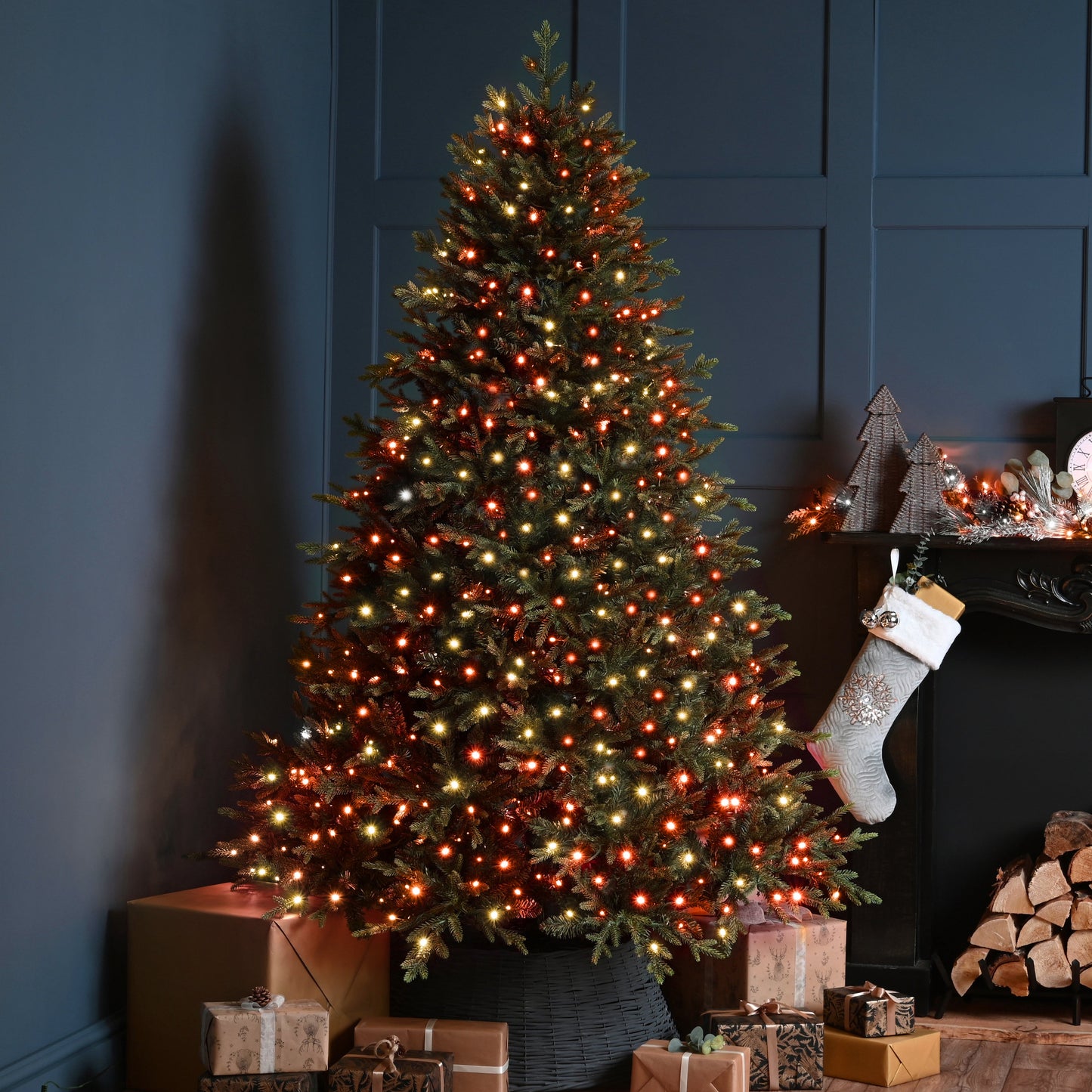 A decorated Christmas tree with warm white and red lights stands beside a fireplace mantel with a hanging stocking, wrapped gifts, and stacked firewood in a dark blue room.