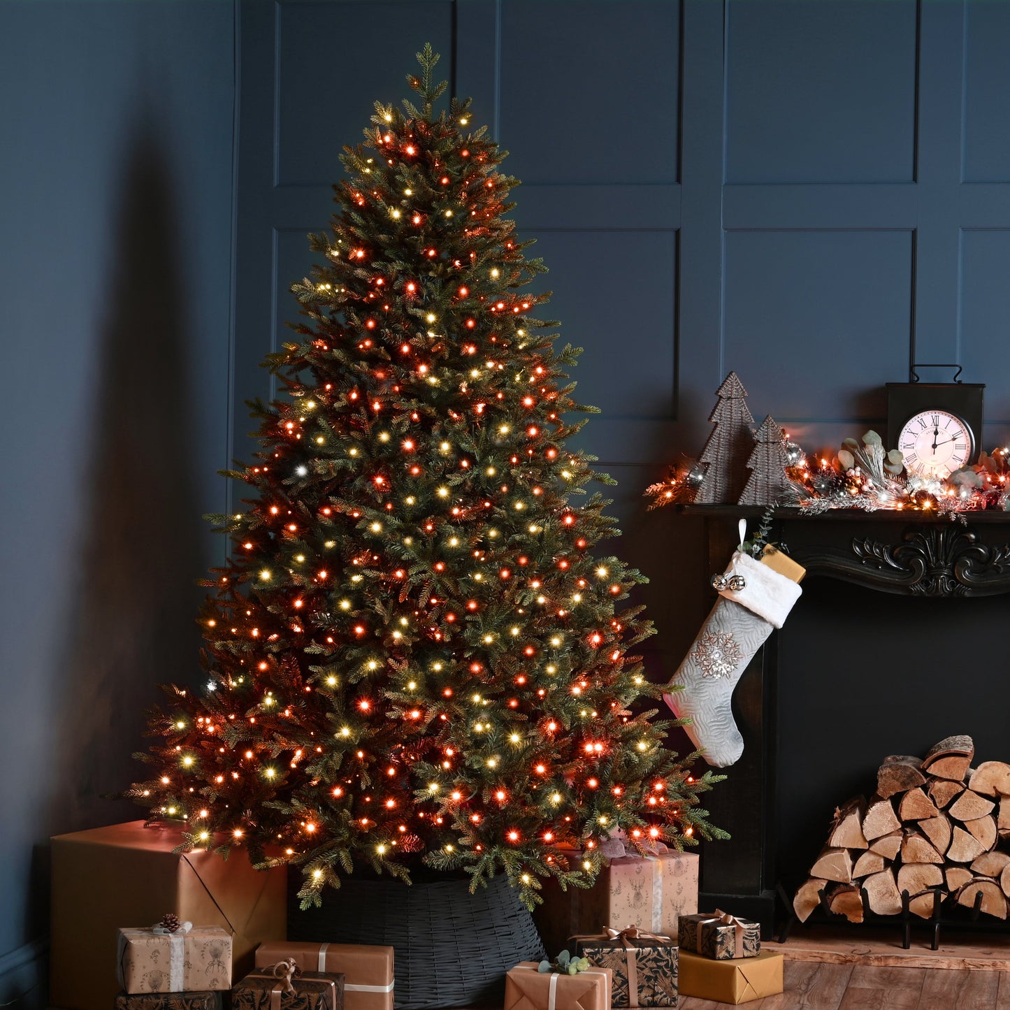 A decorated Christmas tree with warm white and orange lights stands beside a fireplace mantel with a hanging stocking, wrapped gifts, and stacked firewood in a dark blue room.