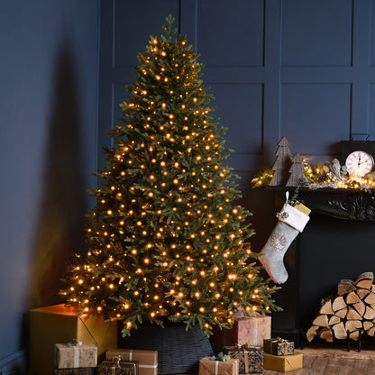 A lit Christmas tree with warm white lights stands beside a dark fireplace mantel decorated with a stocking, small ornaments, and wrapped gifts in a cozy holiday room setting.