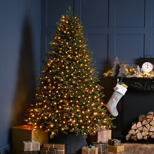 A lit Christmas tree with warm white lights stands beside a dark fireplace mantel decorated with a stocking, small ornaments, and wrapped gifts in a cozy holiday room setting.