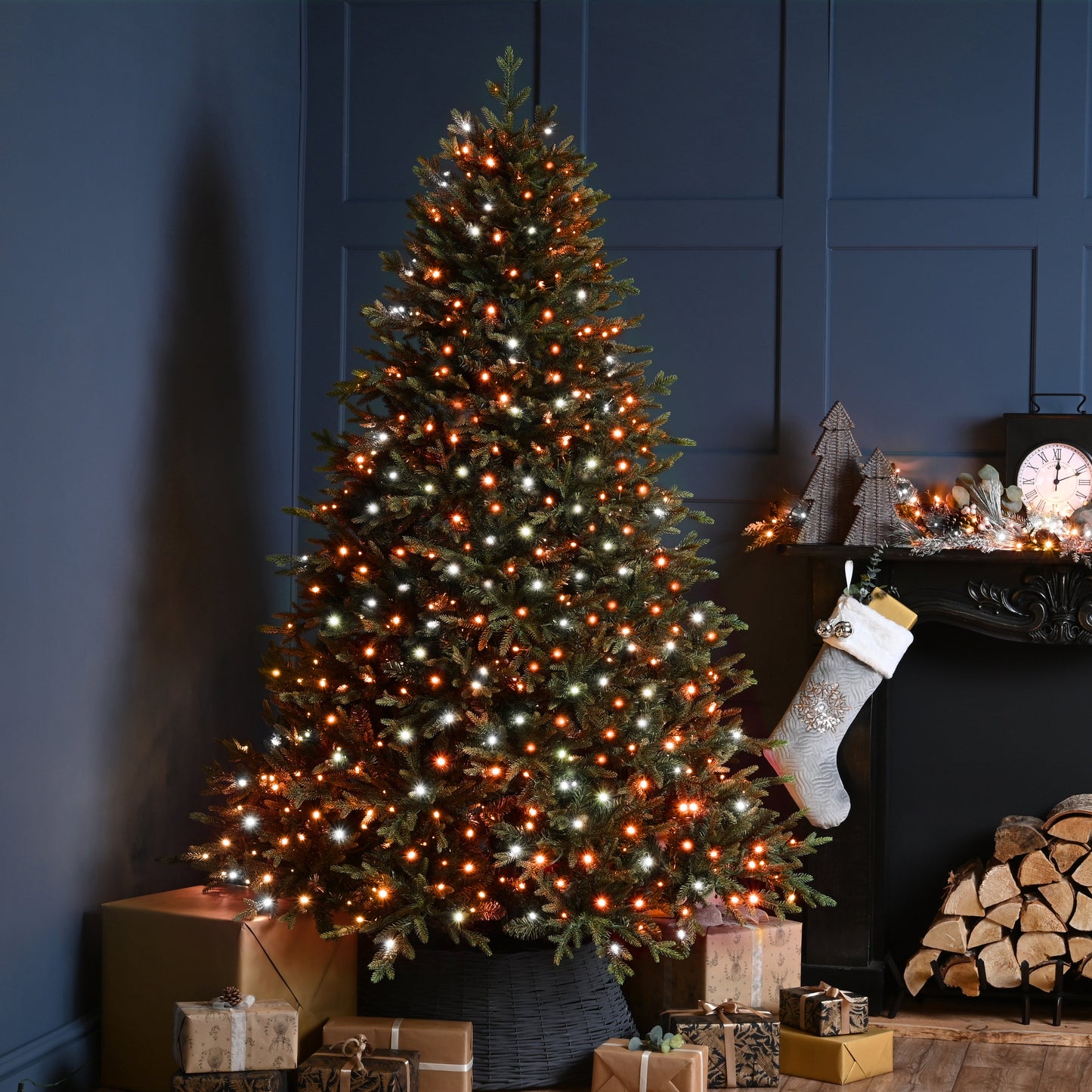 A decorated Christmas tree with red and white lights stands beside a fireplace mantel with a hanging stocking, wrapped gifts, and stacked firewood in a dark blue room.