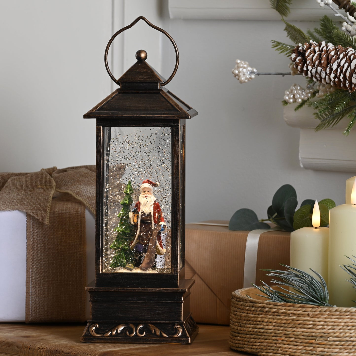 A dark bronze lantern-style Christmas snow globe sits on a wooden table, surrounded by wrapped gifts, lit cream candles, and holiday greenery with pinecones in a softly lit indoor setting.