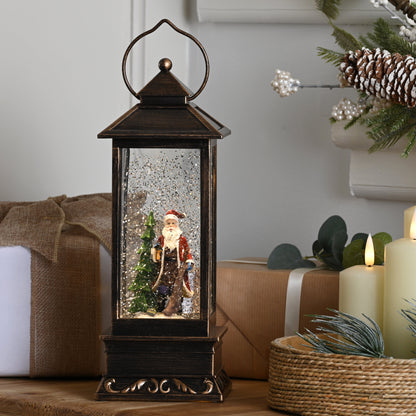 A dark bronze lantern-style Christmas snow globe sits on a wooden table, surrounded by wrapped gifts, lit cream candles, and holiday greenery with pinecones in a softly lit indoor setting.