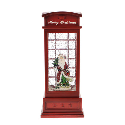 A Christmas-themed red telephone booth decoration with a glittery window, featuring a Santa Claus figurine holding a small tree inside. The top of the booth has 'Merry Christmas' written in white script.