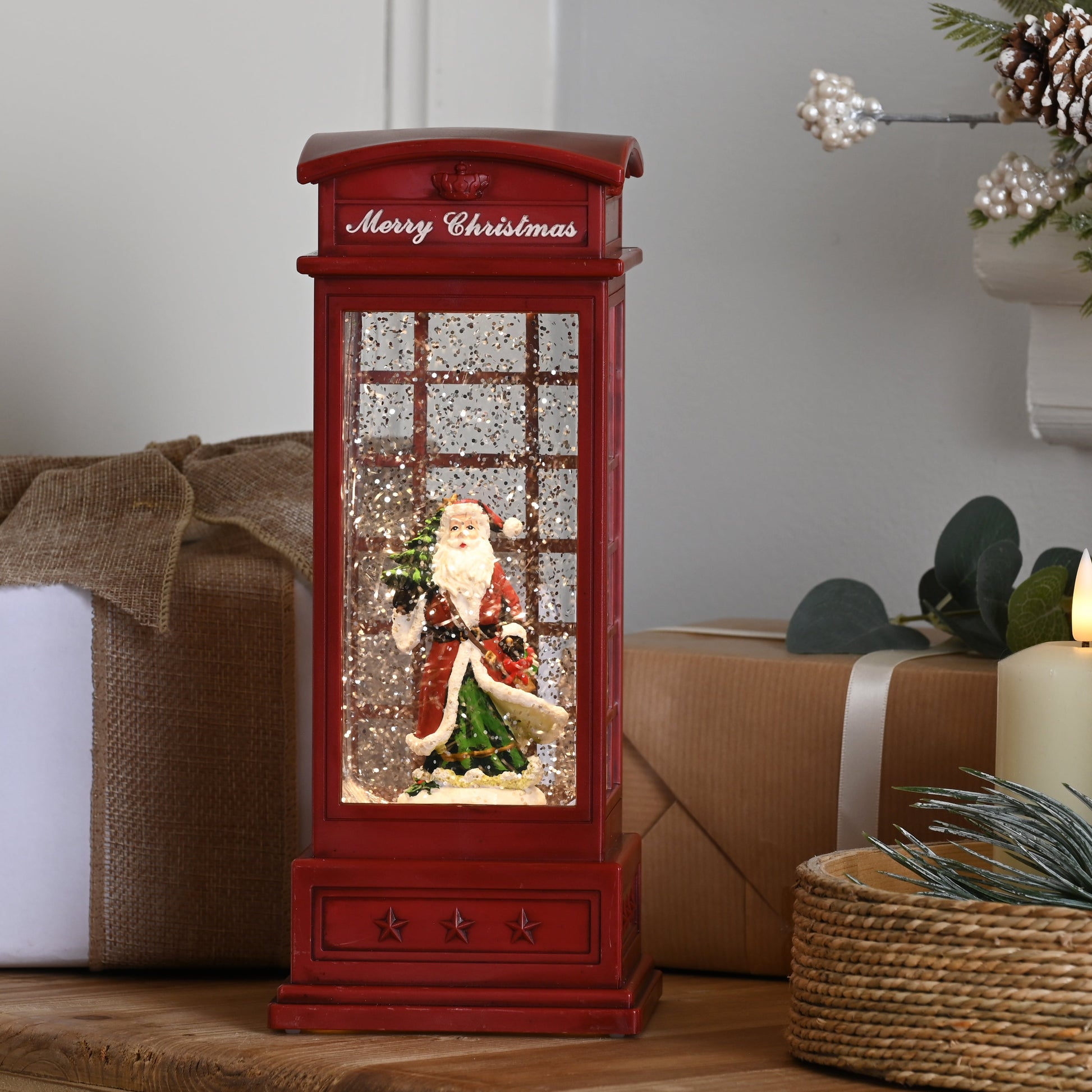 A red telephone booth-shaped Christmas snow globe figurine with Santa Claus inside, styled on a wooden table next to wrapped gifts, lit candles, and a pine garland.