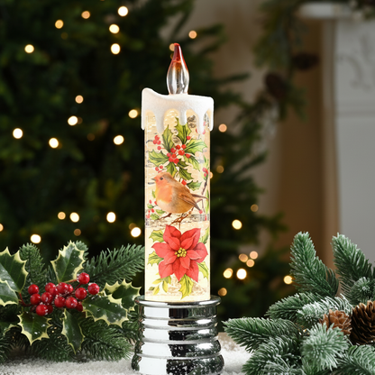 A tall white pillar candle with a Christmas robin and poinsettia design, displayed on a silver base with a blurred Christmas tree and holiday greenery in the background.