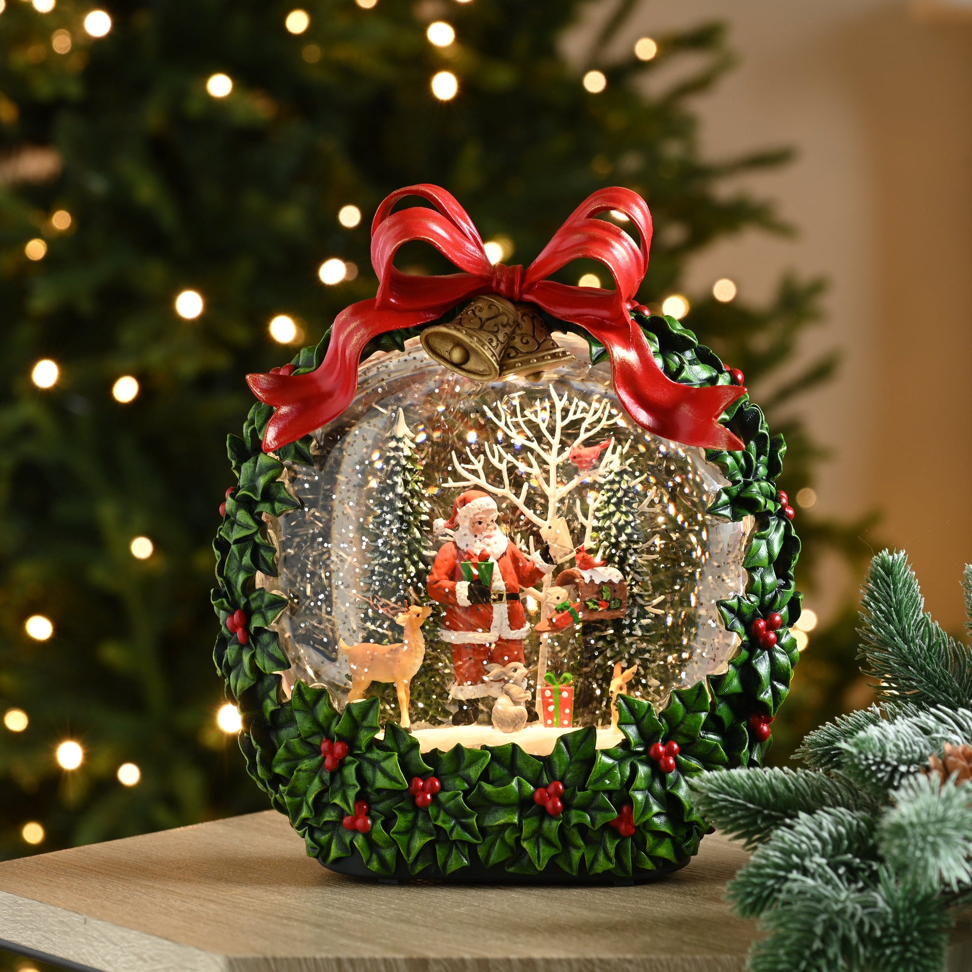 A festive Christmas snow globe decoration displayed on a wooden table in a holiday setting. The ornament features a green wreath base with red berries, a large red ribbon bow, and a clear globe containing a miniature Santa Claus scene with a tree, deer, and warm glowing lights.