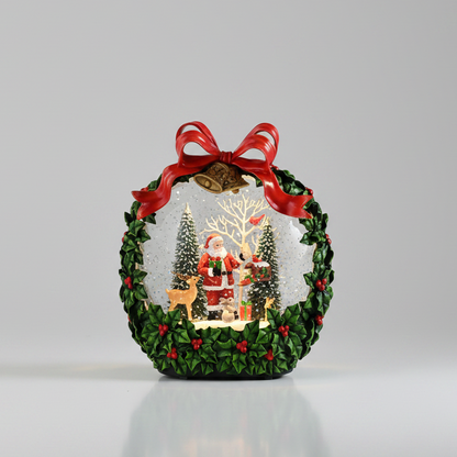 A decorative Christmas snow globe ornament featuring a green wreath border with red berries, a red ribbon bow, and a miniature festive scene inside with Santa, trees, reindeer, and warm lights, photographed on a clean light gray background.