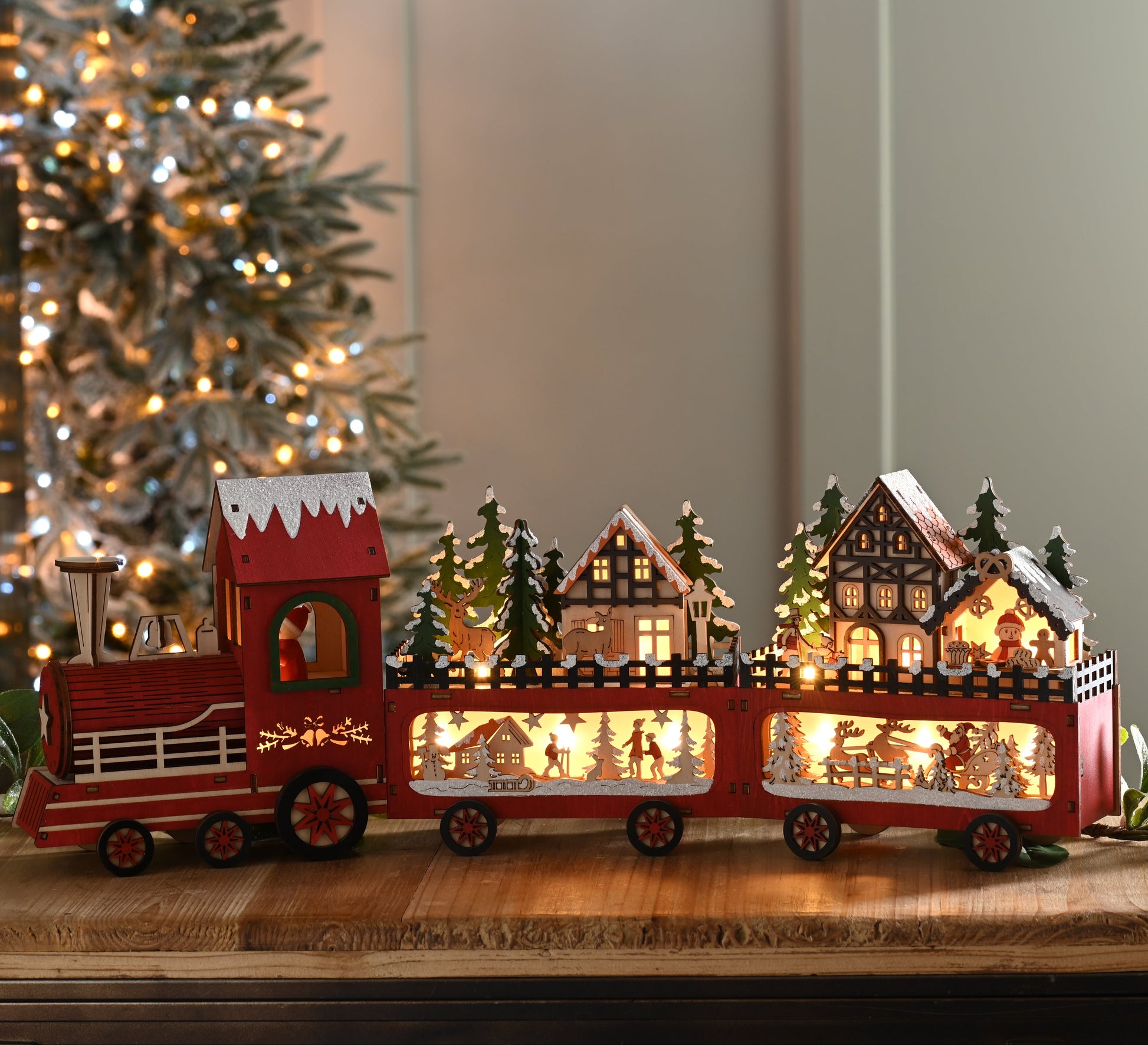 A red wooden Christmas train decoration with illuminated windows, displayed on a wooden surface in front of a softly blurred Christmas tree.