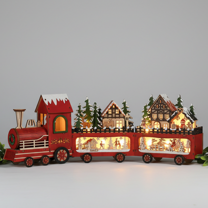 A red wooden Christmas train set with illuminated windows, featuring miniature houses, trees, and festive figures inside the carriages, photographed against a plain light gray studio background.