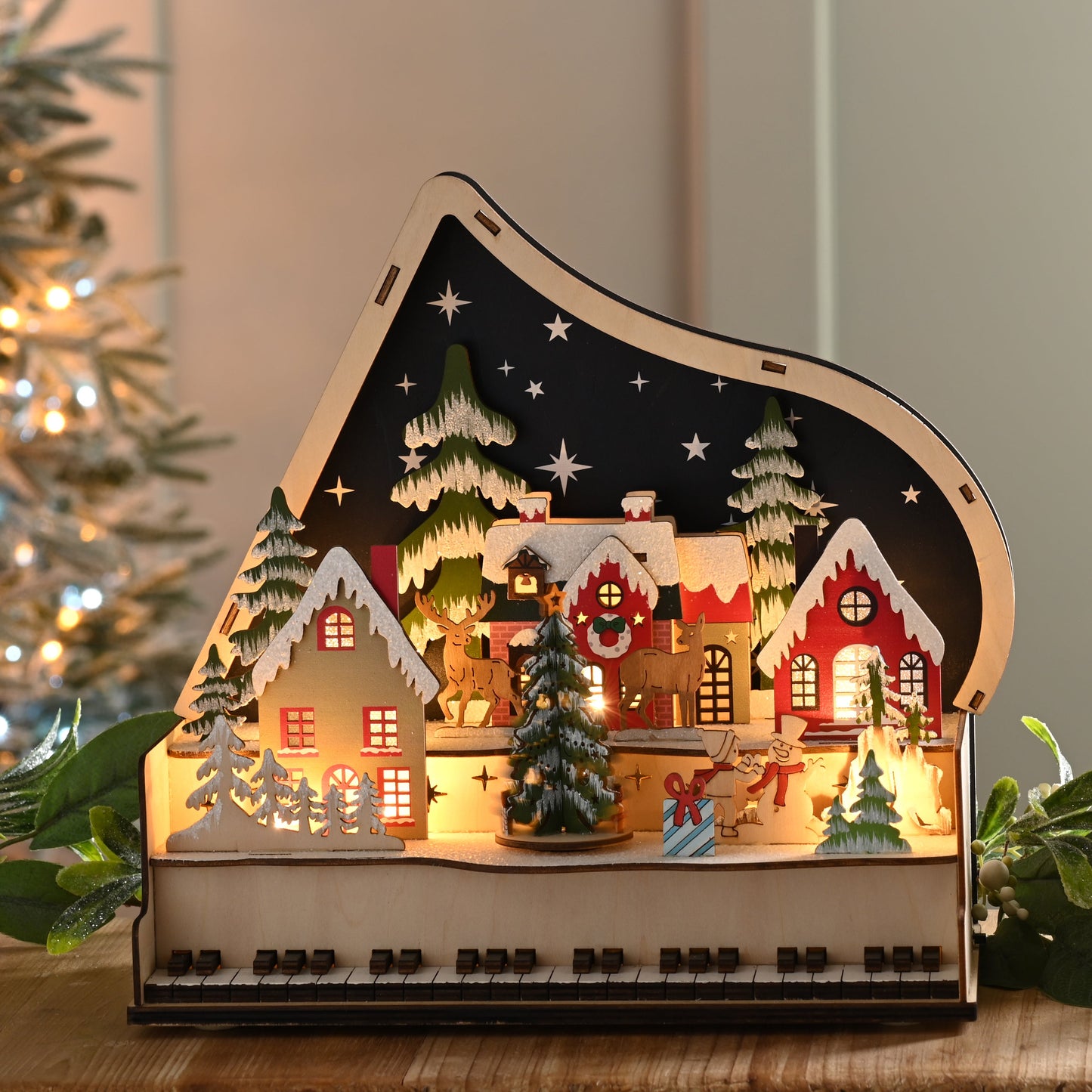 A wooden grand piano-shaped Christmas village night light sits on a wooden table, surrounded by greenery and candles, with a blurred lit Christmas tree in the background.