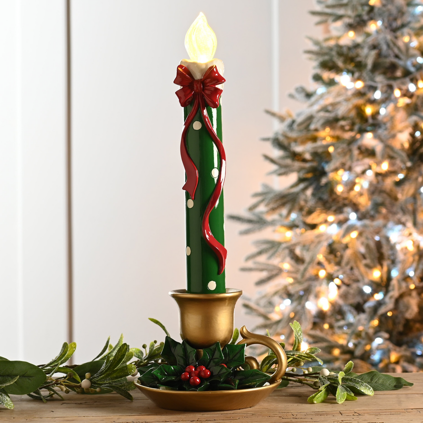 A decorative Christmas candle in a gold-toned holder with a green candle body, red bow and ribbon, white dots, and a lit flame top, styled on a tabletop with holiday greenery and a softly blurred Christmas tree in the background.