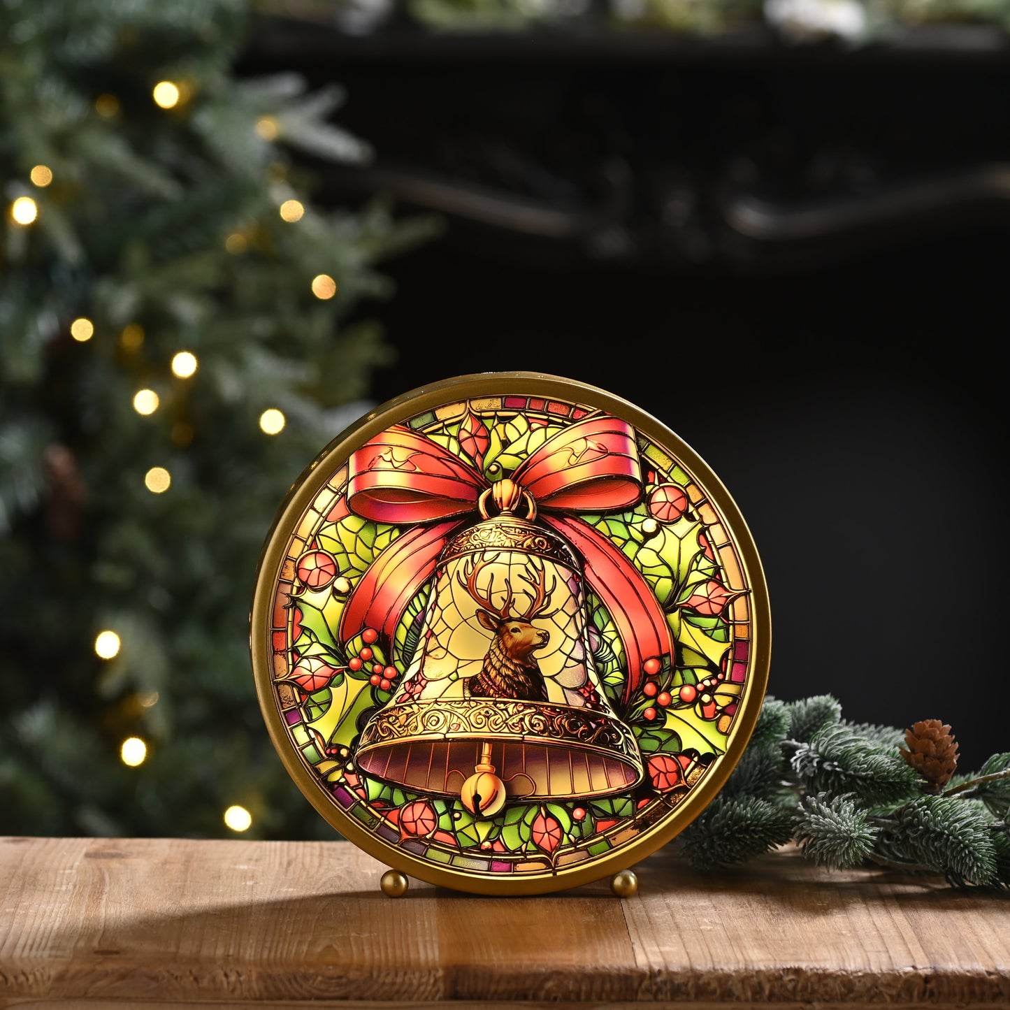 A round decorative Christmas ornament or plaque featuring a gold bell and red bow, displayed on a wooden surface with a blurred Christmas tree and warm lights in the background.
