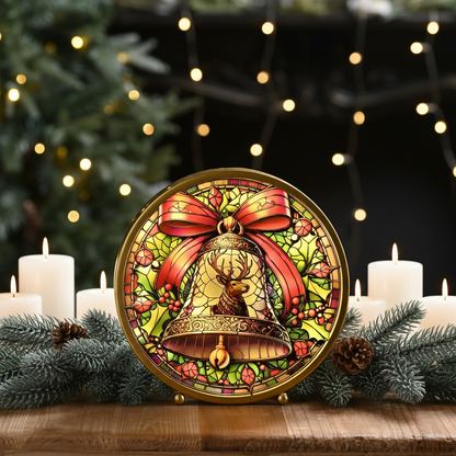 A round decorative Christmas ornament or sign featuring a stained-glass-style design with a golden bell, red bow, green leaves, and pink berries, displayed on a wooden surface with candles, pine branches, and blurred holiday lights in the background.