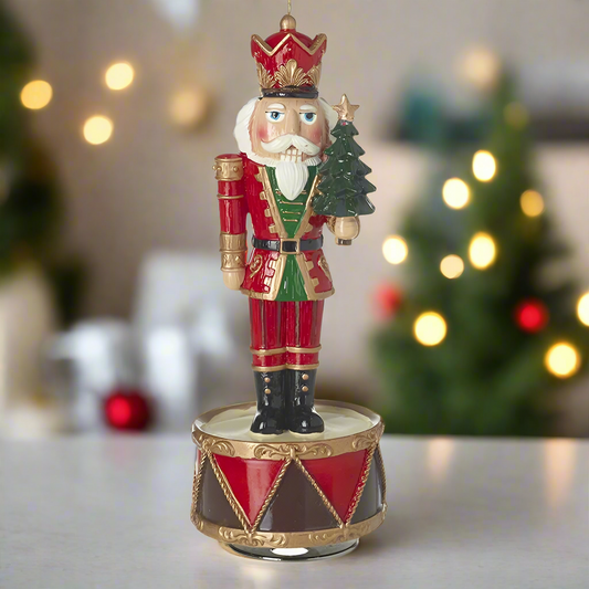 A decorative Christmas music box featuring a vintage style nutcracker holding a tree, standing on a base that resembles a drum. The nutcracker is dressed in red and green, and the base is adorned with gold trim.