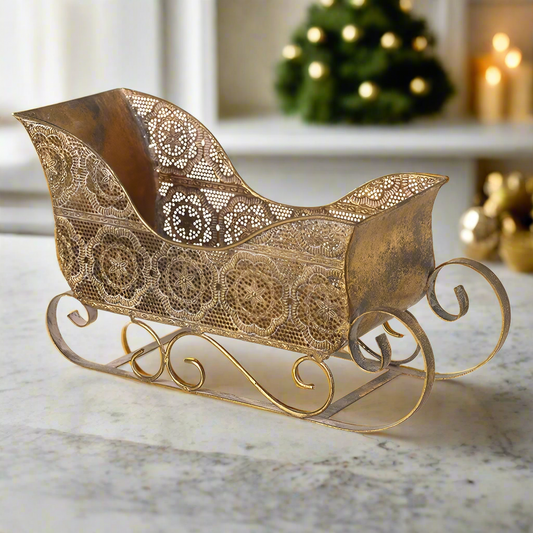 A vintage style gold sleigh decoration placed on a surface with a Christmas tree and lights in the background, indicating a festive setting.