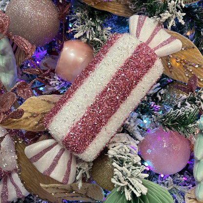A pink candy stripe hanging decoration with glitter and tinsel, surrounded by holiday ornaments and Christmas trees in the background.