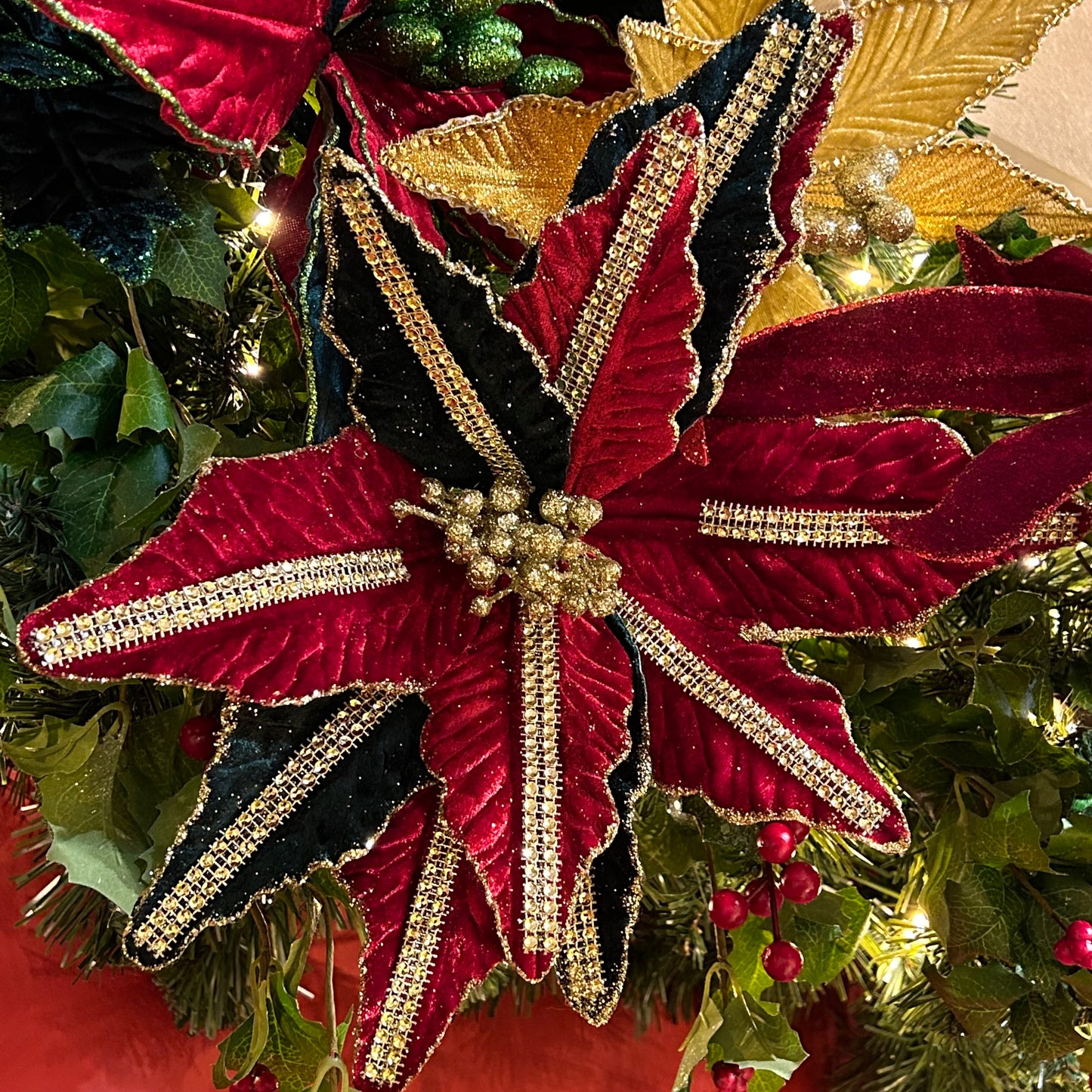A close-up of a red and green artificial poinsettia decoration with gold sequin details along the stem, set against a festive background.
