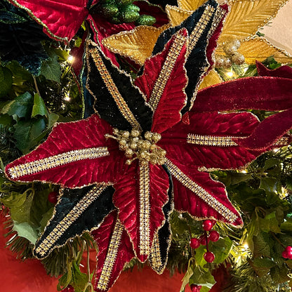 A close-up of a red and green artificial poinsettia decoration with gold sequin details along the stem, set against a festive background.
