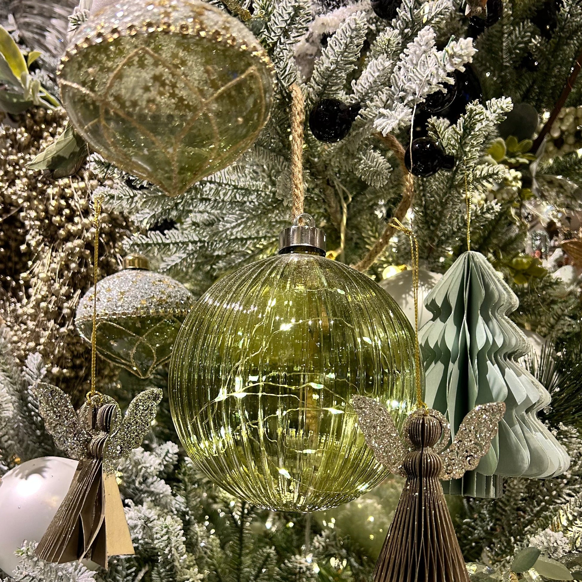A green glass ball Christmas decoration with warm white micro LED lights, hanging from a jute rope, displayed among other festive decorations.