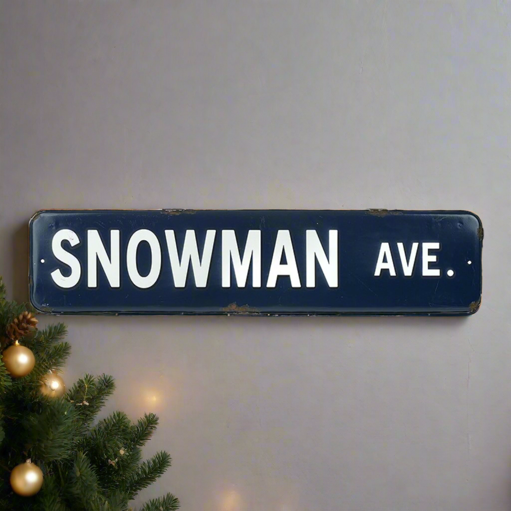 A blue Christmas sign with the words 'Snowman Ave' in white font, placed against a neutral background with a Christmas tree and decorations in the peripheral.
