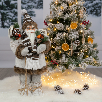 A festive Christmas decoration set featuring a Santa Claus figure in a white and brown knit outfit holding pine branches, next to a decorated flocked Christmas tree with warm string lights and dried orange slices, all arranged on a white faux-fur surface.