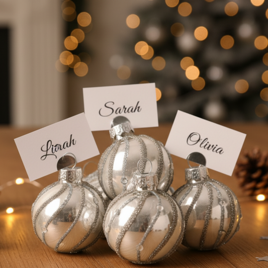 Five silver Christmas baubles with glittery stripes sit on a wooden table, each holding a white name card with a script name (Lilah, Sarah, Olivia). The background is a softly blurred holiday scene with warm bokeh lights.