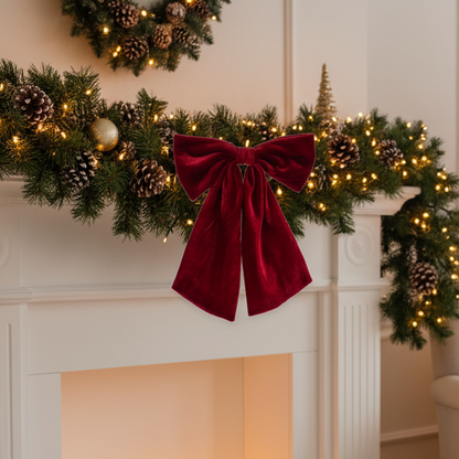 A festive Christmas garland draped over a white fireplace mantel, decorated with pinecones, warm white string lights, a gold ornament, and a large deep red velvet bow in the center.