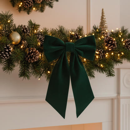 A dark green velvet Christmas bow with long tails, attached to a pine garland decorated with pinecones, a gold ornament, and warm white string lights, hanging above a white fireplace mantel.