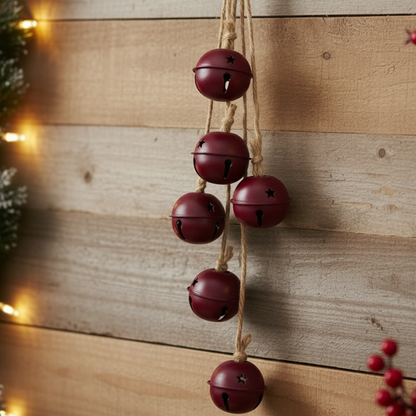 A hanging set of five burgundy jingle bells on a rope, displayed against a rustic wooden wall with warm holiday lights and greenery.