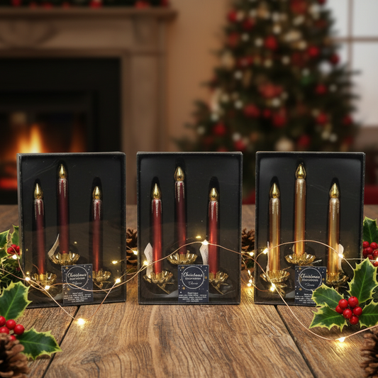 Three boxed sets of Christmas glass taper candles displayed on a wooden table, with a blurred fireplace and decorated Christmas tree in the background. The candles are shown in black gift boxes with gold-toned holders and labels.