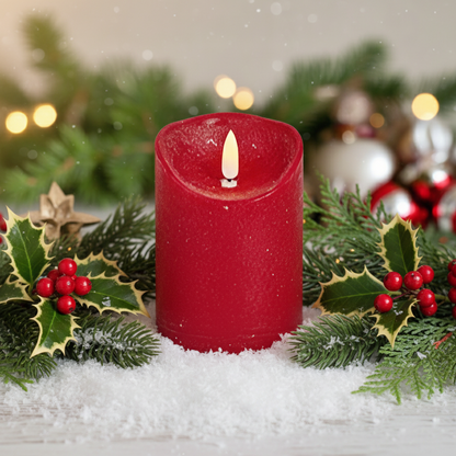 A red cylindrical LED candle with a textured effect, sitting on a surface with Christmas decorations in the background, including a tree and presents.