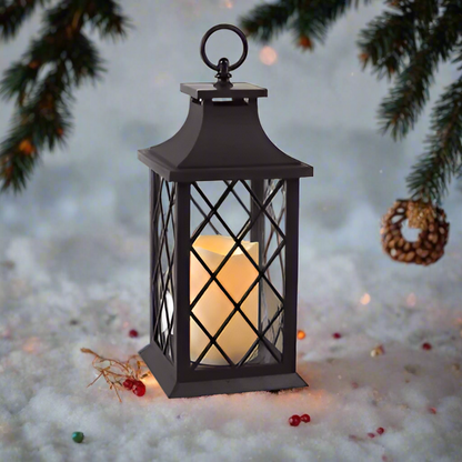 A vintage-style lantern with a flickering candle, primarily black in color, placed on snow-covered ground with Christmas decorations around.