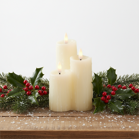 Three white candles with a holly leaf and berry garland on a wooden surface.