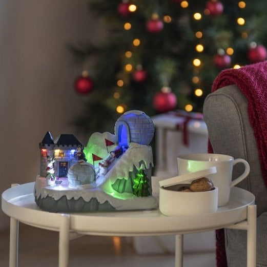 A festive Christmas decoration featuring a mechanical penguin ski slope, with luminous parts and a snowman, set on a table in front of a decorated tree and a cup of coffee.