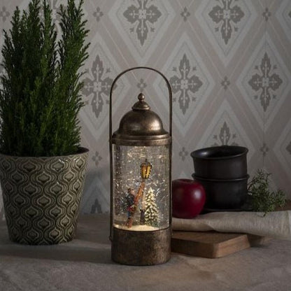 A bronze-colored water lantern with a Dickensian scene inside, surrounded by a glittery snowstorm, placed on a table with a green plant and a bowl in the background.