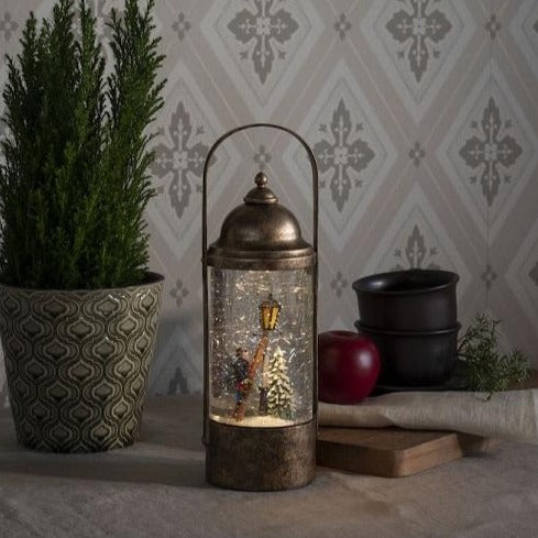 A bronze-colored water lantern with a Dickensian scene inside, surrounded by a glittery snowstorm, placed on a table with a green plant and a bowl in the background.