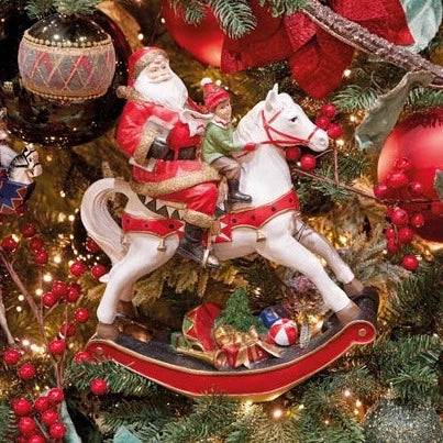 A traditional Christmas decoration featuring Santa Claus and a child on a white rocking horse, with a red saddle and green accents, set against a festive background with holiday ornaments.