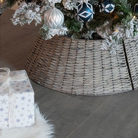 A grey wicker Christmas tree skirt is displayed, covering the base of a decorated Christmas tree and extending out onto the floor, with a white gift box adorned with a snowflake design in front.