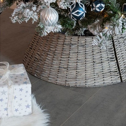 A grey wicker Christmas tree skirt is displayed, covering the base of a decorated Christmas tree and extending out onto the floor, with a white gift box adorned with a snowflake design in front.