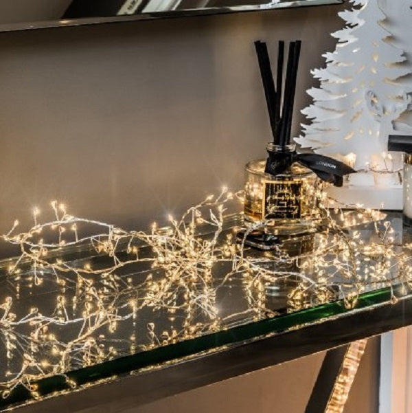 Twinkling LED cluster lights placed on a glass shelf, with a scent diffuser and decorative items in the background, set against a indoor white wall.