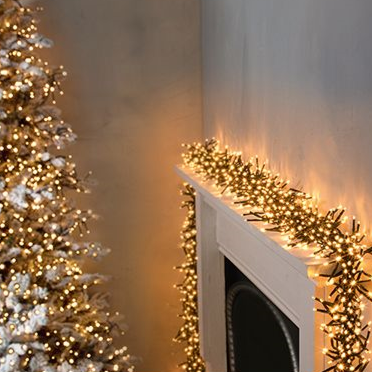 Christmas lights with a vintage gold appearance, clustered together, decorating a fireplace mantle and tree, indicating indoor use.