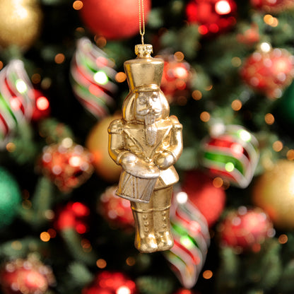 A gold-colored Christmas nutcracker hanging decoration with a uniform, positioned among other holiday decorations on a Christmas tree.