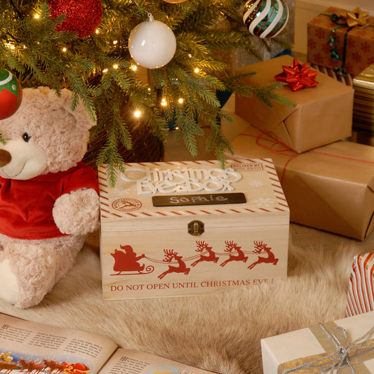 A decorative wooden Christmas Eve box with 'Merry Christmas' text and a reindeer illustration, accompanied by a teddy bear and various gifts in the background.