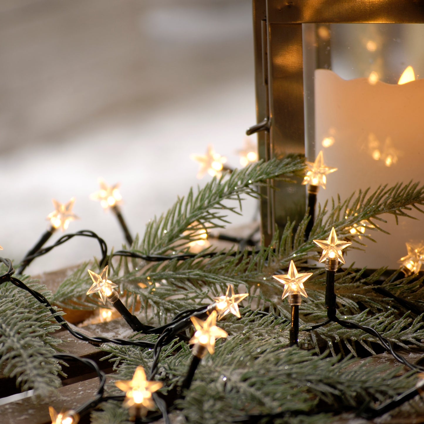 Battery-operated string lights with warm white LEDs, shaped like stars, placed on a green pine branch and in a storage box with snow on the ground.